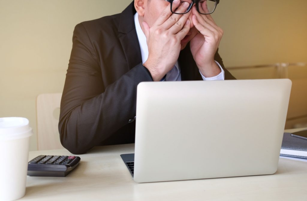 A professional-looking man rubs his eyes under his glasses while taking a break from working on his laptop.