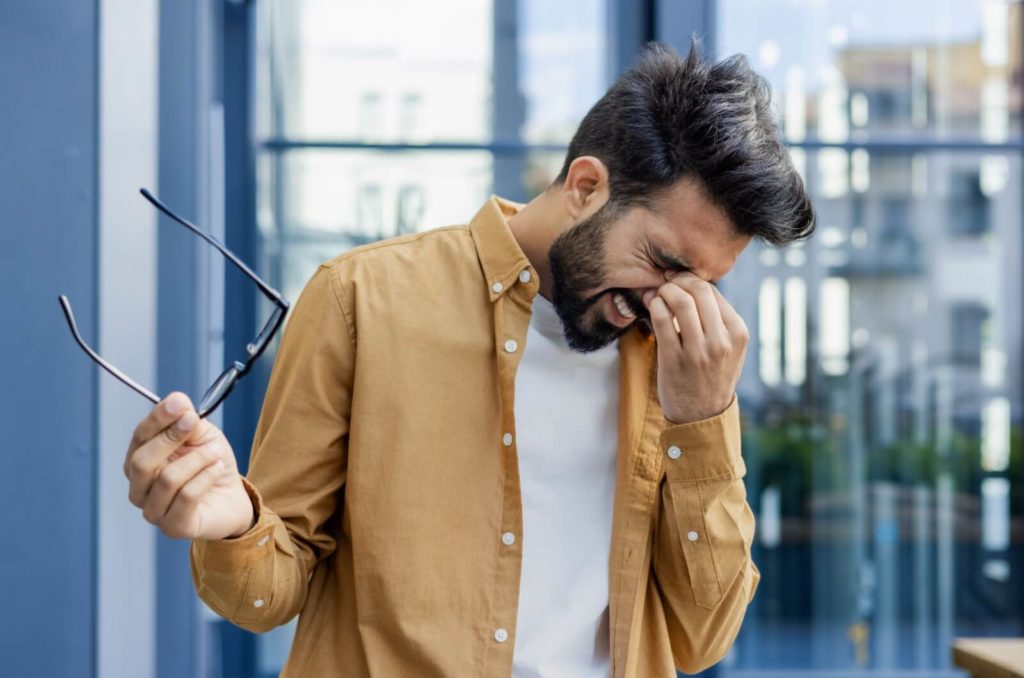 A man holds his eyeglasses in one hand as he pinches the bridge of his nose with the other to try and get some relief from a headache.