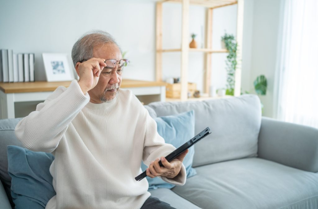 An older man sitting on a couch, adjusting his glasses while trying to read a tablet.