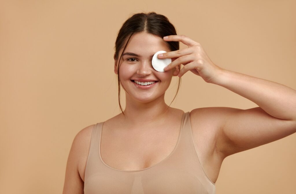 A person in a tan-colored top standing against a tan background and smiling while holding a cotton round up to their left eye.