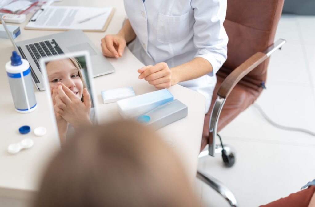 A child carefully inserting a contact lens while looking in the mirror, guided by an optician in a white lab coat. The table holds a laptop and contact lens solution, ready for instruction and support.