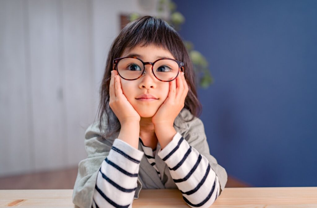 A child with amblyopia, wearing glasses, sitting at a table with their hands resting on their cheeks.
